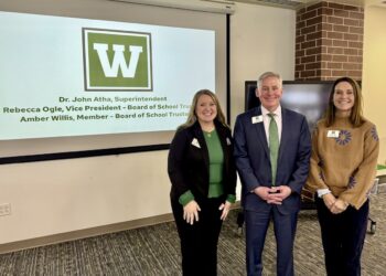 A group of three smiling people—two women and a man—stand together in a modern meeting room. The woman on the left is wearing a black blazer over a green top, the man in the center is in a dark blue suit with a green tie, and the woman on the right is wearing a brown patterned sweater. They are positioned in front of a large projection screen displaying a green box with a white "W" logo and the text: "Dr. John Atha, Superintendent," "Rebecca Ogle, Vice President - Board of School Trustees," and "Amber Willis, Member - Board of School Trustees." A dark brick pillar and a large television monitor are visible to the right.