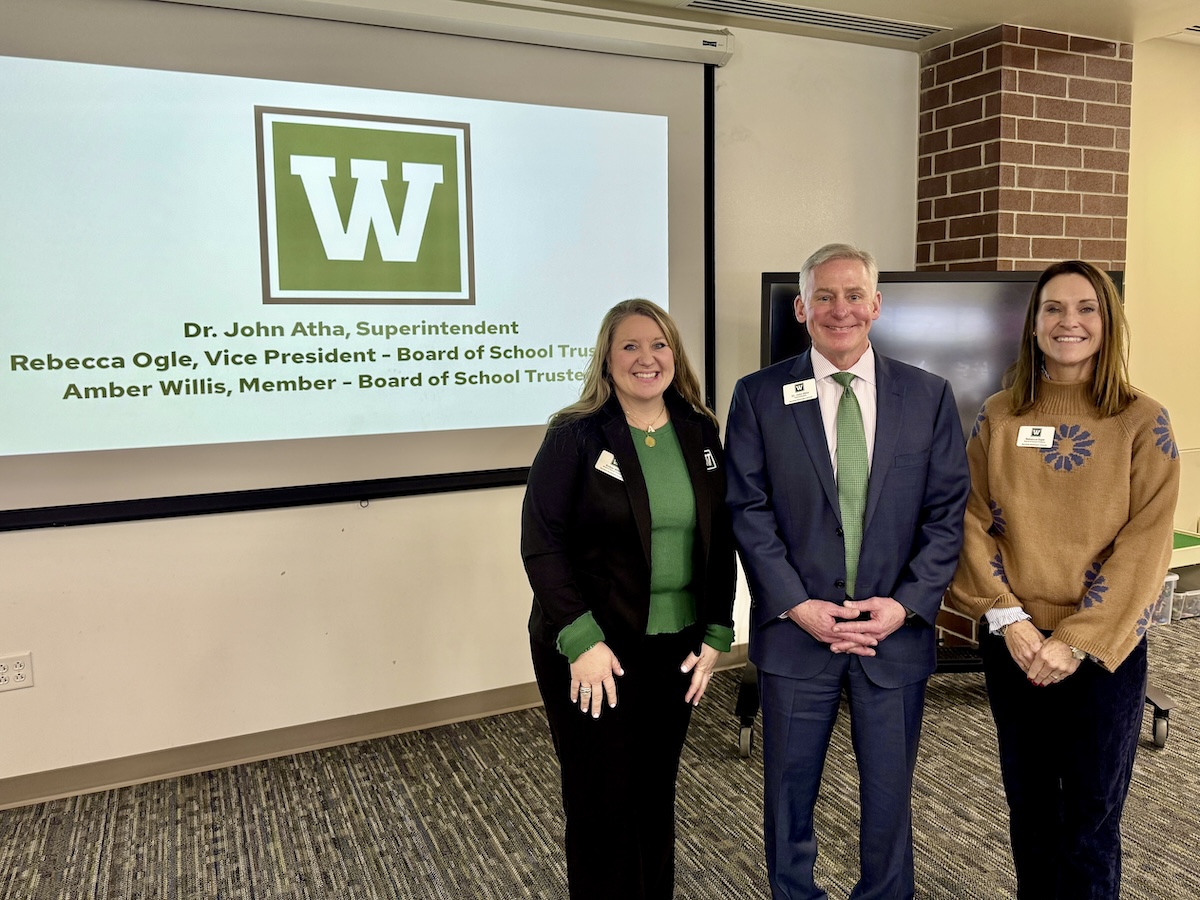 A group of three smiling people—two women and a man—stand together in a modern meeting room. The woman on the left is wearing a black blazer over a green top, the man in the center is in a dark blue suit with a green tie, and the woman on the right is wearing a brown patterned sweater. They are positioned in front of a large projection screen displaying a green box with a white "W" logo and the text: "Dr. John Atha, Superintendent," "Rebecca Ogle, Vice President - Board of School Trustees," and "Amber Willis, Member - Board of School Trustees." A dark brick pillar and a large television monitor are visible to the right.