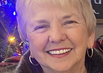 A close-up portrait of a smiling woman with short, blonde hair and gold hoop earrings. She wears a grey top and a light-colored jacket with a dark, fuzzy collar. To her left, a decorated Christmas tree with blue, orange, and white lights is partially visible. In the upper right corner, a chalkboard sign with the name "Alyona" written in white cursive is also partially visible.