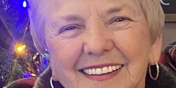 A close-up portrait of a smiling woman with short, blonde hair and gold hoop earrings. She wears a grey top and a light-colored jacket with a dark, fuzzy collar. To her left, a decorated Christmas tree with blue, orange, and white lights is partially visible. In the upper right corner, a chalkboard sign with the name "Alyona" written in white cursive is also partially visible.