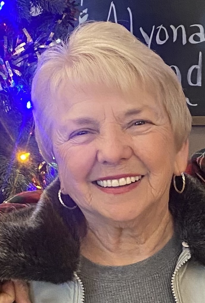 A close-up portrait of a smiling woman with short, blonde hair and gold hoop earrings. She wears a grey top and a light-colored jacket with a dark, fuzzy collar. To her left, a decorated Christmas tree with blue, orange, and white lights is partially visible. In the upper right corner, a chalkboard sign with the name "Alyona" written in white cursive is also partially visible.