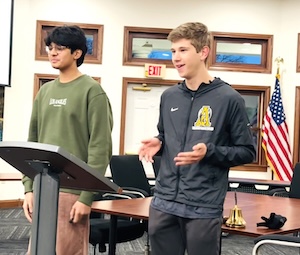 Two teenage boys stand at a podium in a formal meeting room, likely a school board or community meeting. The boy on the right, wearing a black Nike zip-up jacket with a yellow and white school emblem, is speaking and gesturing with his hands. To his left, another boy in an olive green sweatshirt looks on. Behind them is a large wooden conference table with a brass bell, an American flag, and several wood-framed windows.