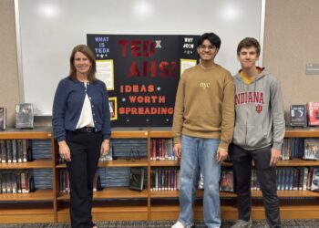 In a library setting, a woman and two teenage boys pose in front of a trifold display board. The woman on the left wears a blue cardigan and black slacks. The boy in the center wears a tan sweatshirt and jeans, and the boy on the right wears a grey Indiana University hoodie. The black display board behind them features red and yellow text that reads "TEDx AHS! IDEAS WORTH SPREADING." Wooden bookshelves filled with books line the wall behind them.
