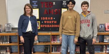In a library setting, a woman and two teenage boys pose in front of a trifold display board. The woman on the left wears a blue cardigan and black slacks. The boy in the center wears a tan sweatshirt and jeans, and the boy on the right wears a grey Indiana University hoodie. The black display board behind them features red and yellow text that reads "TEDx AHS! IDEAS WORTH SPREADING." Wooden bookshelves filled with books line the wall behind them.