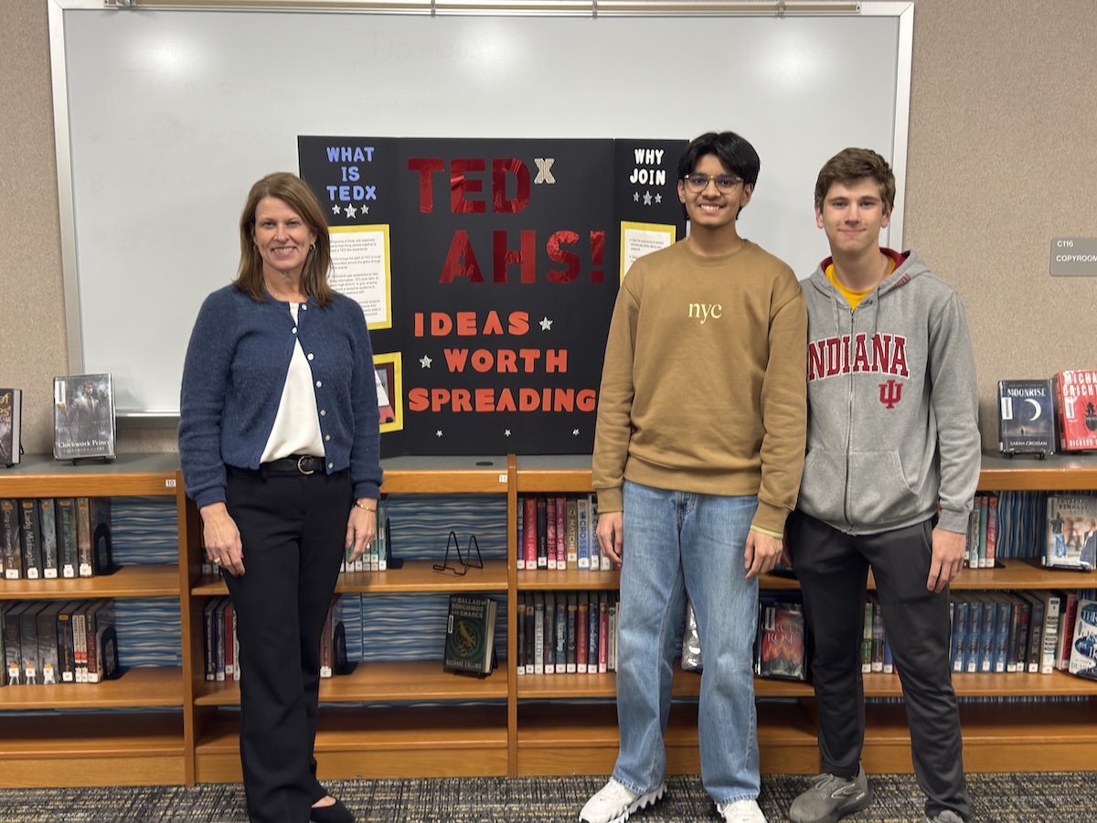 In a library setting, a woman and two teenage boys pose in front of a trifold display board. The woman on the left wears a blue cardigan and black slacks. The boy in the center wears a tan sweatshirt and jeans, and the boy on the right wears a grey Indiana University hoodie. The black display board behind them features red and yellow text that reads "TEDx AHS! IDEAS WORTH SPREADING." Wooden bookshelves filled with books line the wall behind them.