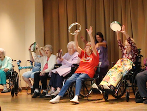 A group of elderly people in wheelchairs sit in a row on a stage, joyfully participating in a musical activity. Several individuals are enthusiastically raising their arms, holding and shaking tambourines. A younger woman stands behind them, smiling and assisting. The background consists of a simple white wall on the left and a large, tan-colored curtain across the rest of the stage.