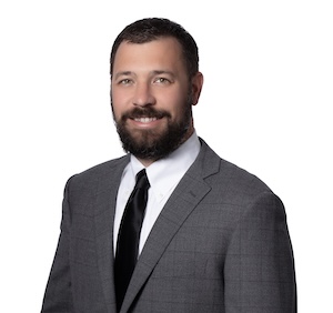 A professional headshot of a smiling man with a full dark beard and short hair. He is wearing a grey patterned suit jacket, a white collared shirt, and a solid black tie, set against a plain white background.