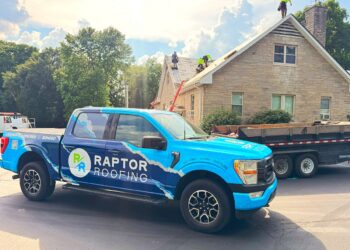A bright blue Ford F-150 pickup truck with a large "RAPTOR ROOFING" wrap is parked on a street in front of a tan stone house. In the background, a crew of roofers in neon safety vests is actively working on the gabled roof, which is partially stripped down to the wooden decking. A large black dumpster trailer is parked next to the truck, and a red ladder is leaned against the side of the house under a bright, sunny sky with puffy white clouds