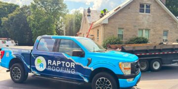 A bright blue Ford F-150 pickup truck with a large "RAPTOR ROOFING" wrap is parked on a street in front of a tan stone house. In the background, a crew of roofers in neon safety vests is actively working on the gabled roof, which is partially stripped down to the wooden decking. A large black dumpster trailer is parked next to the truck, and a red ladder is leaned against the side of the house under a bright, sunny sky with puffy white clouds
