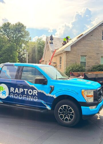 A bright blue Ford F-150 pickup truck with a large "RAPTOR ROOFING" wrap is parked on a street in front of a tan stone house. In the background, a crew of roofers in neon safety vests is actively working on the gabled roof, which is partially stripped down to the wooden decking. A large black dumpster trailer is parked next to the truck, and a red ladder is leaned against the side of the house under a bright, sunny sky with puffy white clouds