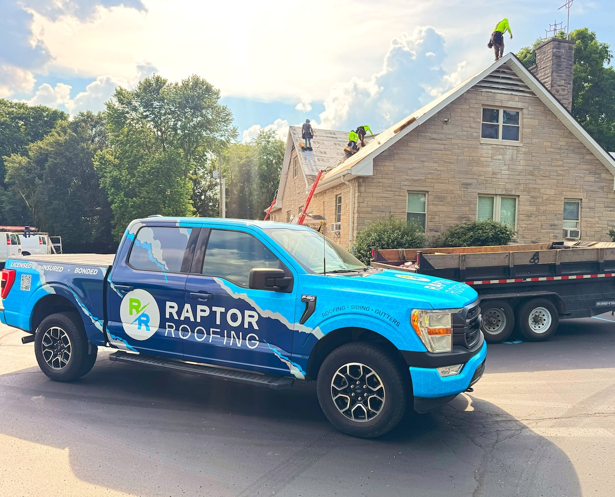 A bright blue Ford F-150 pickup truck with a large "RAPTOR ROOFING" wrap is parked on a street in front of a tan stone house. In the background, a crew of roofers in neon safety vests is actively working on the gabled roof, which is partially stripped down to the wooden decking. A large black dumpster trailer is parked next to the truck, and a red ladder is leaned against the side of the house under a bright, sunny sky with puffy white clouds