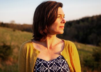 A woman with shoulder-length brown hair looks off to the side, bathed in the warm, golden light of a sunset. She wears a yellow cardigan over a blue and white patterned top. The background shows a soft-focus landscape of rolling hills and trees under a clear sky.
