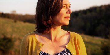A woman with shoulder-length brown hair looks off to the side, bathed in the warm, golden light of a sunset. She wears a yellow cardigan over a blue and white patterned top. The background shows a soft-focus landscape of rolling hills and trees under a clear sky.