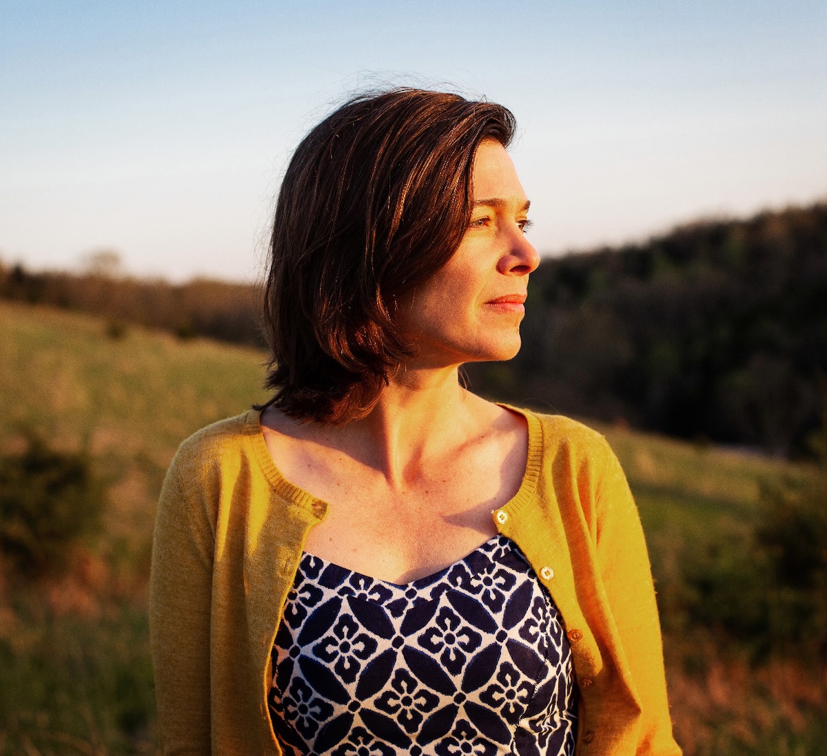 A woman with shoulder-length brown hair looks off to the side, bathed in the warm, golden light of a sunset. She wears a yellow cardigan over a blue and white patterned top. The background shows a soft-focus landscape of rolling hills and trees under a clear sky.
