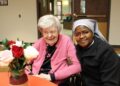 An elderly woman in a pink cardigan and a Catholic nun in a grey and black habit smile warmly together at a table. The elderly woman is laughing joyfully, while the nun has her arm around her in a supportive gesture. In the foreground, a vase of red and white roses sits on the orange-clothed table.