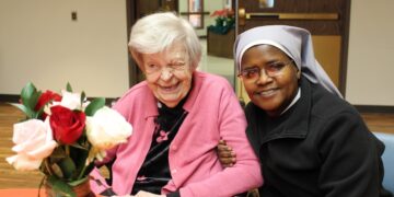 An elderly woman in a pink cardigan and a Catholic nun in a grey and black habit smile warmly together at a table. The elderly woman is laughing joyfully, while the nun has her arm around her in a supportive gesture. In the foreground, a vase of red and white roses sits on the orange-clothed table.