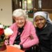 An elderly woman in a pink cardigan and a Catholic nun in a grey and black habit smile warmly together at a table. The elderly woman is laughing joyfully, while the nun has her arm around her in a supportive gesture. In the foreground, a vase of red and white roses sits on the orange-clothed table.
