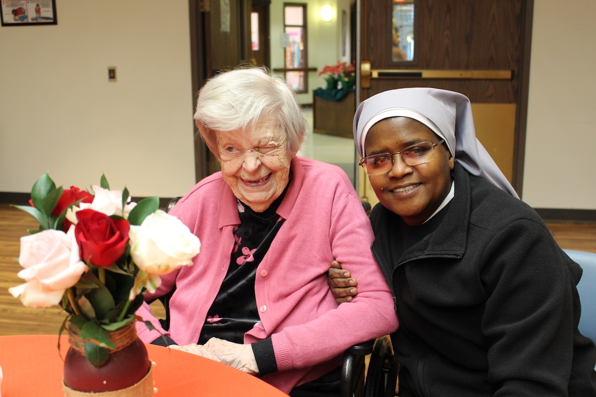 An elderly woman in a pink cardigan and a Catholic nun in a grey and black habit smile warmly together at a table. The elderly woman is laughing joyfully, while the nun has her arm around her in a supportive gesture. In the foreground, a vase of red and white roses sits on the orange-clothed table.