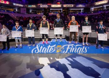 A wide shot of the 190-pound weight class podium at the 2026 IHSAA Boys Wrestling State Finals inside Gainbridge Fieldhouse in Indianapolis. Eight state-placing wrestlers stand on a multi-tiered blue podium decorated with the "IHSAA State Finals" logo. The state champion, Michael White of Lawrence North (undefeated at 47-0), stands on the highest central tier wearing a gold medal. Other place-winners stand to his sides, each holding a large white bracket sheet and wearing their respective medals. The background shows the brightly lit arena with spectators in the stands and various corporate sponsor banners for Salesforce, IndyStar, and FanDuel.