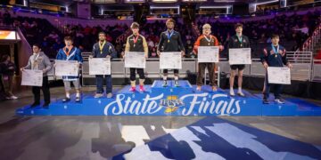 A wide shot of the 190-pound weight class podium at the 2026 IHSAA Boys Wrestling State Finals inside Gainbridge Fieldhouse in Indianapolis. Eight state-placing wrestlers stand on a multi-tiered blue podium decorated with the "IHSAA State Finals" logo. The state champion, Michael White of Lawrence North (undefeated at 47-0), stands on the highest central tier wearing a gold medal. Other place-winners stand to his sides, each holding a large white bracket sheet and wearing their respective medals. The background shows the brightly lit arena with spectators in the stands and various corporate sponsor banners for Salesforce, IndyStar, and FanDuel.