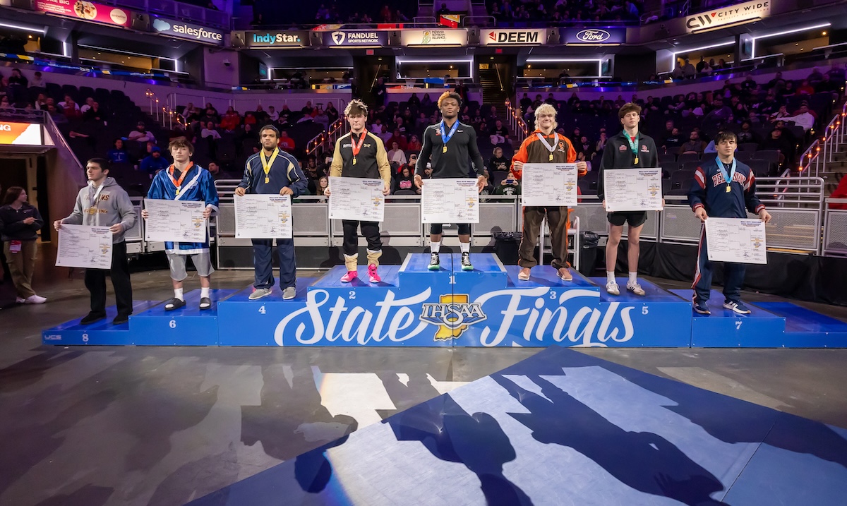 A wide shot of the 190-pound weight class podium at the 2026 IHSAA Boys Wrestling State Finals inside Gainbridge Fieldhouse in Indianapolis. Eight state-placing wrestlers stand on a multi-tiered blue podium decorated with the "IHSAA State Finals" logo. The state champion, Michael White of Lawrence North (undefeated at 47-0), stands on the highest central tier wearing a gold medal. Other place-winners stand to his sides, each holding a large white bracket sheet and wearing their respective medals. The background shows the brightly lit arena with spectators in the stands and various corporate sponsor banners for Salesforce, IndyStar, and FanDuel.
