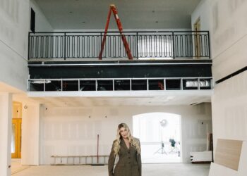 A full-length portrait of a woman standing in the center of a spacious, unfinished interior at Westwood Vowery & Estate, a brand-new wedding venue in Westfield, Indiana. She is smiling confidently and wearing a dark brown, patterned power suit with black heels. The large room is in the final stages of construction, with exposed drywall panels on the high ceilings and walls. Directly behind her is a black metal balcony railing for a second-story mezzanine, where a red ladder is perched. The space features large arched doorways and a clean, light-colored concrete floor, hinting at the venue's future modern-meets-traditional aesthetic.