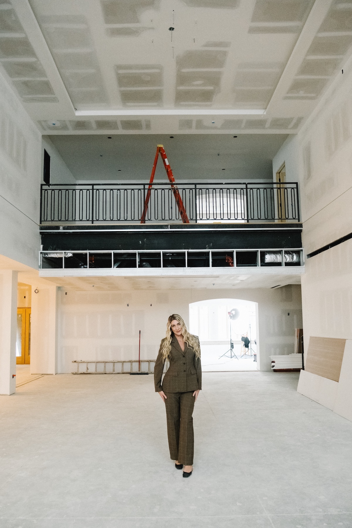 A full-length portrait of a woman standing in the center of a spacious, unfinished interior at Westwood Vowery & Estate, a brand-new wedding venue in Westfield, Indiana. She is smiling confidently and wearing a dark brown, patterned power suit with black heels. The large room is in the final stages of construction, with exposed drywall panels on the high ceilings and walls. Directly behind her is a black metal balcony railing for a second-story mezzanine, where a red ladder is perched. The space features large arched doorways and a clean, light-colored concrete floor, hinting at the venue's future modern-meets-traditional aesthetic.