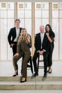 A professional group portrait of the Westwood Vowery & Estate ownership team posing on the stone steps of their new venue in Westfield, Indiana. The four individuals are dressed in sophisticated, formal attire. On the left, Seth Dempsey (Hospitality Director) stands in a black suit and white shirt, with his hand on the shoulder of his wife, Shelby Dempsey (Founder, Owner, and COO), who is seated on a black stool. Shelby is wearing a textured brown plaid power suit and black heels. To their right, Chris Henry (Partner) is seated on a matching stool in a black suit and light-colored tie, while his wife, Tana Henry (Partner), stands behind him in a long black dress with her hand on his shoulder. They are positioned in front of tall, modern windows with delicate gold framing that overlook the estate's grounds.