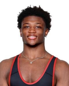 A professional studio headshot of Michael White, a state-champion wrestler from Lawrence North High School. He is a young Black man with short, textured hair and a warm, confident smile. He is wearing a black wrestling singlet with red trim, small silver stud earrings, and a thin silver chain necklace with a small circular pendant. The background is a solid, clean white, focusing the attention on his friendly and determined expression.