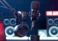 A professional podcast or radio studio setup featuring a large black condenser microphone on a boom arm and a pair of over-ear headphones hanging nearby. In the blurred background, a glowing red "ON AIR" sign sits atop studio monitor speakers, and a coffee mug rests on the desk in the foreground. The room is lit with moody blue and red atmospheric lighting.