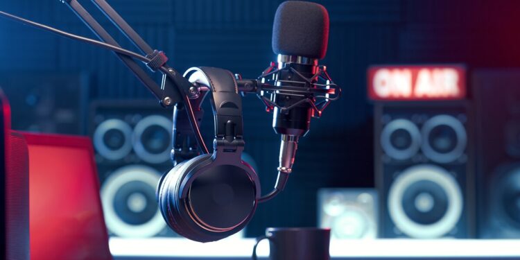 A professional podcast or radio studio setup featuring a large black condenser microphone on a boom arm and a pair of over-ear headphones hanging nearby. In the blurred background, a glowing red "ON AIR" sign sits atop studio monitor speakers, and a coffee mug rests on the desk in the foreground. The room is lit with moody blue and red atmospheric lighting.