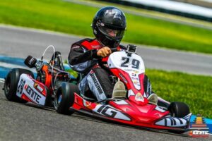A high-action shot of a go-kart racer navigating a paved track. The driver, wearing a sleek black carbon-fiber helmet with a white Bell logo and a red, black, and white racing suit, is firmly gripping the steering wheel of a red and white Margay Ignite kart numbered "29." A small GoPro camera is mounted on the front fairing. The kart is equipped with a black racing engine on the right and features "IGNITE" and "Margay" branding on its side pods. The background shows a vibrant green grassy field and a gray track surface under bright daylight.