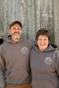 A man and woman standing side-by-side, smiling in front of a rustic, grey wooden barn wall. They are both wearing matching dark grey hoodies with a circular logo on the chest that reads "Shrock Family Farm." The man also wears a brown baseball cap.