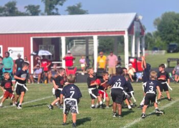 A dynamic shot of a youth flag football game in progress on a lush green field. Several young players are captured in action; a boy in a black "Warriors" jersey clutches a red football while being pursued by players in navy blue jerseys featuring a white "CP" logo. In the background, a red building with a white-pillared porch provides shade for spectators. The scene is bright and sunny, highlighting the energy of the local community sports event.