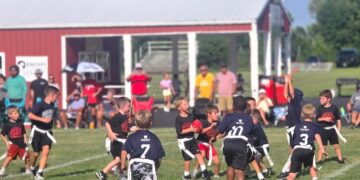 A dynamic shot of a youth flag football game in progress on a lush green field. Several young players are captured in action; a boy in a black "Warriors" jersey clutches a red football while being pursued by players in navy blue jerseys featuring a white "CP" logo. In the background, a red building with a white-pillared porch provides shade for spectators. The scene is bright and sunny, highlighting the energy of the local community sports event.