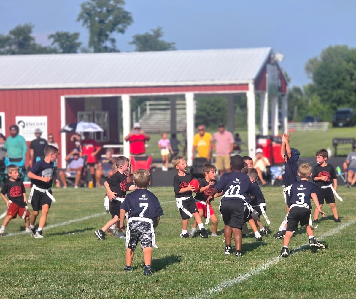 A dynamic shot of a youth flag football game in progress on a lush green field. Several young players are captured in action; a boy in a black "Warriors" jersey clutches a red football while being pursued by players in navy blue jerseys featuring a white "CP" logo. In the background, a red building with a white-pillared porch provides shade for spectators. The scene is bright and sunny, highlighting the energy of the local community sports event.