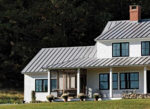 A modern white farmhouse with a striking grey standing-seam metal roof and black-trimmed windows. The home features a cozy screened-in porch on the left, leading to an open porch area with outdoor seating and a low stone wall. Four potted white chrysanthemums line the porch edge, and a red brick chimney rises from the roofline. The house is set against a backdrop of dense, dark green trees and a grassy lawn in the foreground.