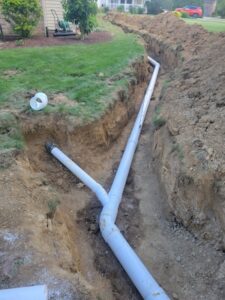 A newly installed white PVC sewer line sits in a deep, narrow trench in a grassy backyard. The main pipe runs straight down the length of the trench, with a smaller branch pipe connecting into it at a Y-junction. Large mounds of excavated dirt line the right side of the trench, and a well-maintained lawn and a house are visible in the background.