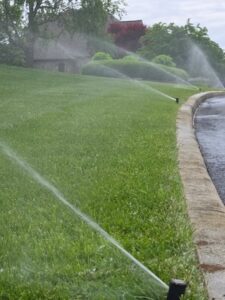 Several active sprinkler heads along a stone-lined driveway spray water in long, steady streams across a lush, green, sloping lawn. In the background, a residential home is partially visible behind manicured bushes and trees under an overcast sky.
