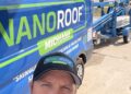 A selfie of a man with a beard wearing a black NanoRoof Michiana baseball cap. He is standing in front of a blue company van with a large NanoRoof logo and the slogan "Extend your roof life 10, 15, or 20 years!" A blue Genie trailer-mounted boom lift is visible in the background on a sunny day.