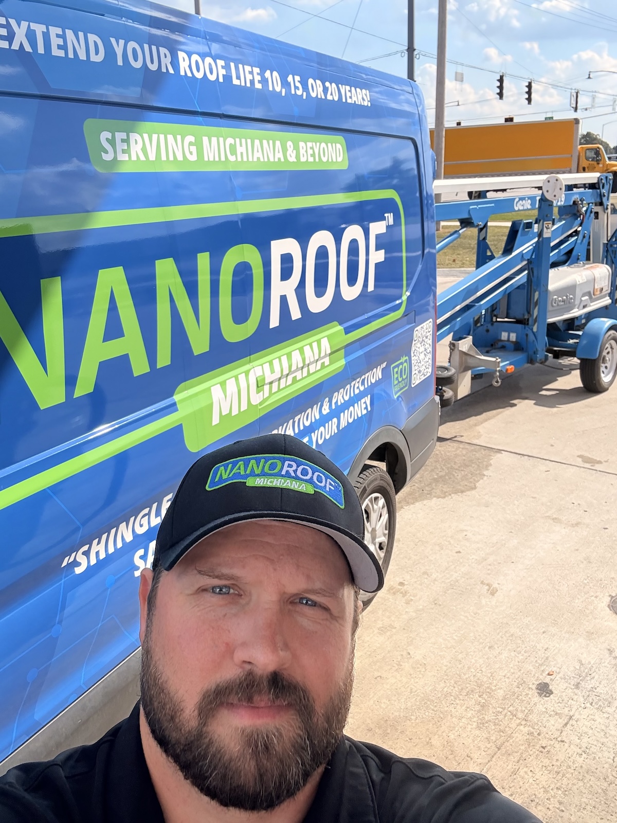 A selfie of a man with a beard wearing a black NanoRoof Michiana baseball cap. He is standing in front of a blue company van with a large NanoRoof logo and the slogan "Extend your roof life 10, 15, or 20 years!" A blue Genie trailer-mounted boom lift is visible in the background on a sunny day.