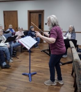 A side-profile shot shows a woman with long, silver hair leading a choir rehearsal. She is wearing a maroon long-sleeved shirt, dark jeans, and tan sneakers, and is holding a thin wooden baton in her right hand. She stands behind a blue music stand holding an open book of sheet music. In the background, several senior women are seated in blue chairs, some holding music folders and looking toward the conductor. The room has light-colored walls, wood-paneled doors, and a polished wood floor.