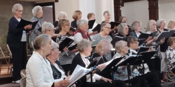 A group of approximately twenty senior women perform as a choir in what appears to be a church or community hall with dark wood paneling and white pillars. The women are arranged in three rows, with the front row seated and the back two rows standing. They are all singing with focused expressions, holding black music folders or binders. Many are dressed in patterned tops—stripes, polka dots, and music-note prints—while others wear solid-colored sweaters and jackets. Several music stands are positioned in the foreground.