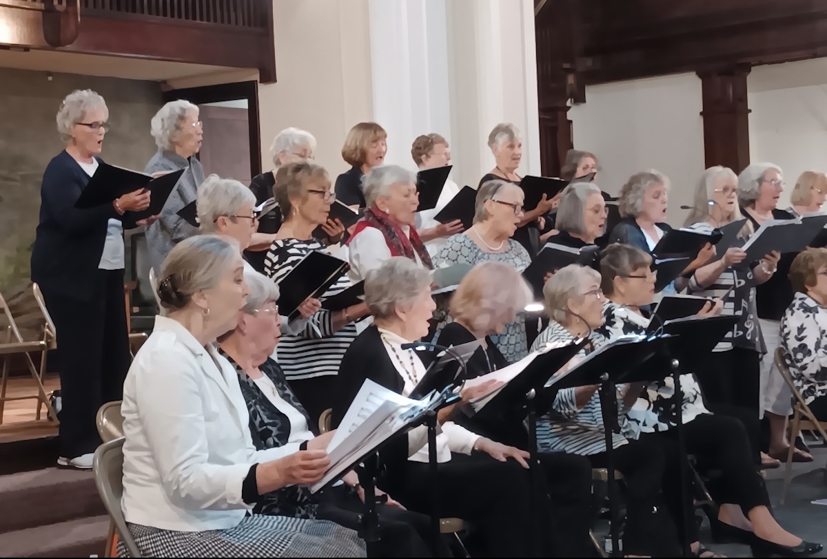 A group of approximately twenty senior women perform as a choir in what appears to be a church or community hall with dark wood paneling and white pillars. The women are arranged in three rows, with the front row seated and the back two rows standing. They are all singing with focused expressions, holding black music folders or binders. Many are dressed in patterned tops—stripes, polka dots, and music-note prints—while others wear solid-colored sweaters and jackets. Several music stands are positioned in the foreground.