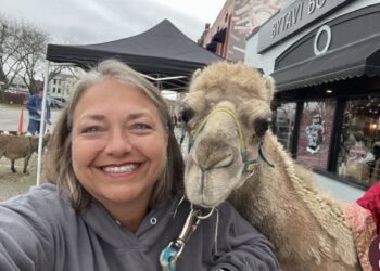 A close-up selfie of a smiling woman with graying hair and blue eyes, wearing a gray hooded sweatshirt. She is posing next to a light brown dromedary camel that is leaning its head toward her. The camel has a blue and yellow halter. In the background, a street scene in downtown Franklin, Indiana, is visible, including a black canopy tent, a brick building with a sign that reads "BYTAVI BOUTIQUE," and a small brown goat.