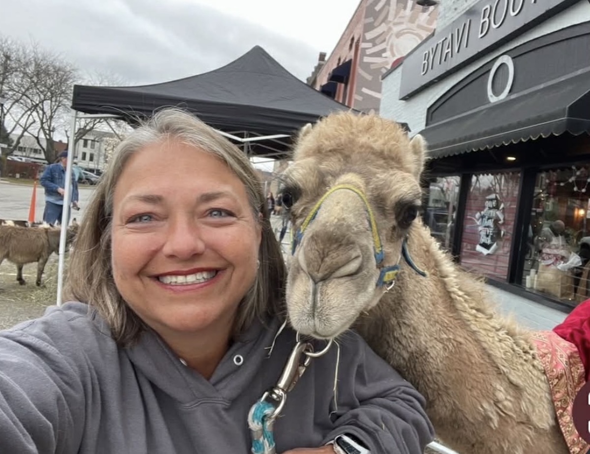 A close-up selfie of a smiling woman with graying hair and blue eyes, wearing a gray hooded sweatshirt. She is posing next to a light brown dromedary camel that is leaning its head toward her. The camel has a blue and yellow halter. In the background, a street scene in downtown Franklin, Indiana, is visible, including a black canopy tent, a brick building with a sign that reads "BYTAVI BOUTIQUE," and a small brown goat.