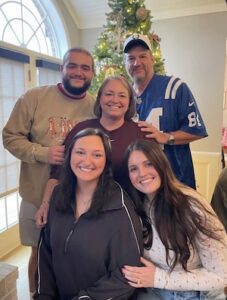 A smiling family of five—two women in the foreground, a woman in the center, and two men standing behind them—pose for a portrait in front of a decorated Christmas tree. The two younger women in the front are seated or kneeling; one wears a black quarter-zip sweatshirt and the other a white long-sleeved top. The woman in the center wears a maroon shirt. Behind her, one man wears a tan sweatshirt and the other is in a blue Indianapolis Colts jersey and a matching baseball cap. The brightly lit room features a large arched window with a white frame in the background.