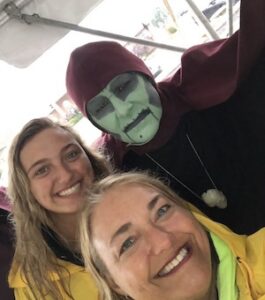 A close-up selfie of two smiling women in yellow raincoats posing with a person in a "Wicked Witch" costume. The woman in the foreground has light hair and green eyes, while the woman behind her has long blonde hair. The person in costume has pale green face paint, dark eyes, and a black line drawn down their chin, wearing a maroon hood and a black top with a large, light-colored pendant. They are all outdoors under a white tent canopy, with a street and buildings visible in the background.