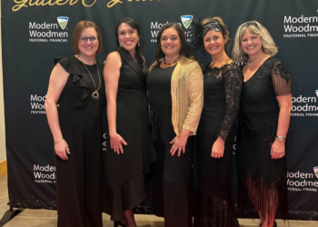 Five women stand together, smiling, on a red carpet in front of a black backdrop at a formal event. They are all dressed in elegant black evening attire, including dresses and jumpsuits. The woman in the center wears a shimmering gold cardigan over her black outfit, and the woman to her right wears a delicate black lace headpiece. The backdrop features the words "Glitter & Gratitude GALA" in large gold script at the top, along with repeated white logos for "Modern Woodmen FRATERNAL FINANCIAL.