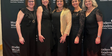 Five women stand together, smiling, on a red carpet in front of a black backdrop at a formal event. They are all dressed in elegant black evening attire, including dresses and jumpsuits. The woman in the center wears a shimmering gold cardigan over her black outfit, and the woman to her right wears a delicate black lace headpiece. The backdrop features the words "Glitter & Gratitude GALA" in large gold script at the top, along with repeated white logos for "Modern Woodmen FRATERNAL FINANCIAL.